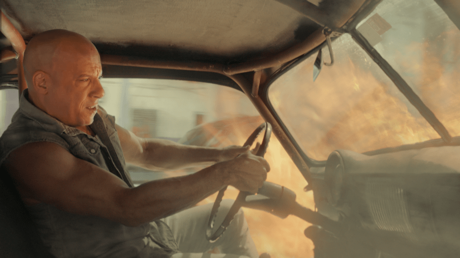 Muscular driver gripping the steering wheel and leaning forward in a roll‑caged off‑road vehicle as a huge explosion/fireball erupts outside, conveying high-speed, high-stress action.